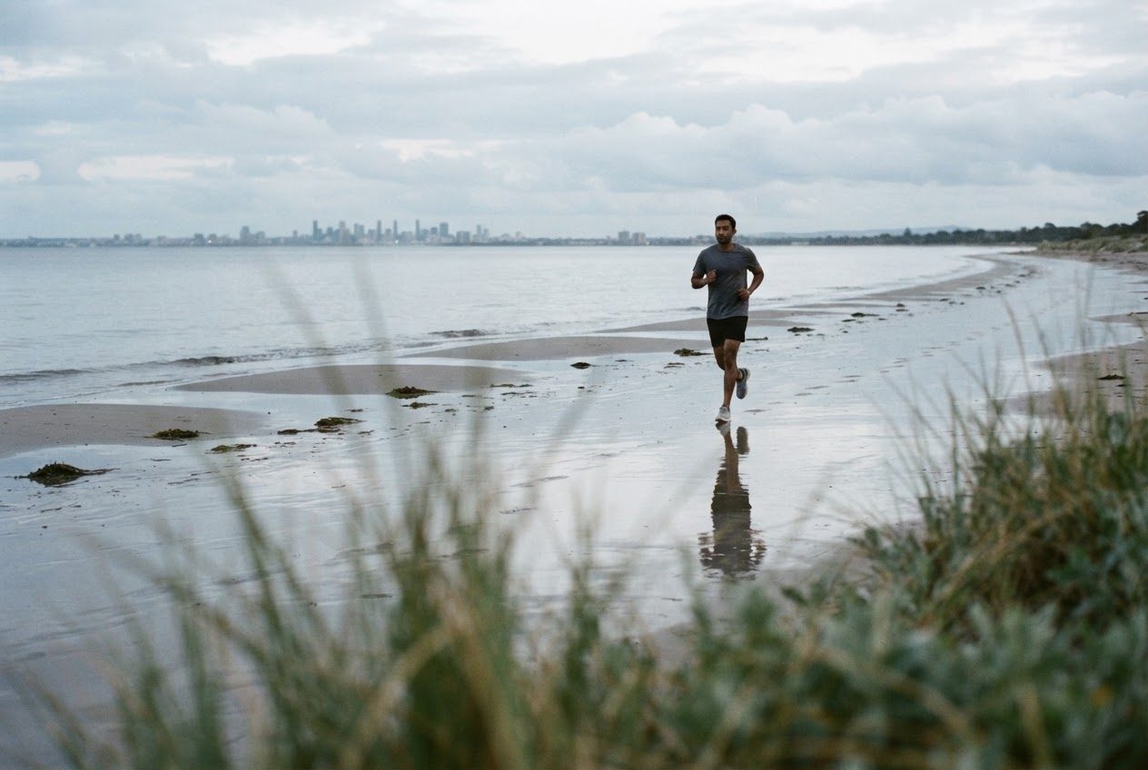 A South Asian man in his thirties, an ordinary beginner runner, follows a quiet path into the distance with the day's conditions clearly visible on firm wet sand at a city beach