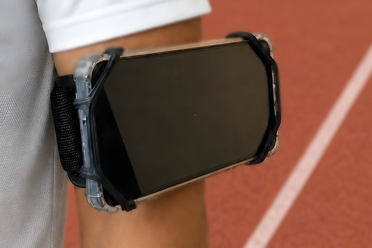 A phone in a clear case sits sideways in a black open corner holder on a black band on an upper arm beside a red running track.
