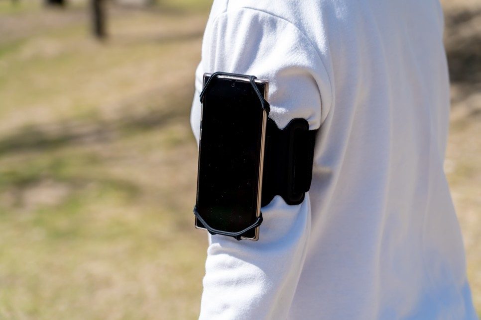 A black armband holding a phone in an open corner holder is worn on the upper arm over a white long sleeve in a sunny park.