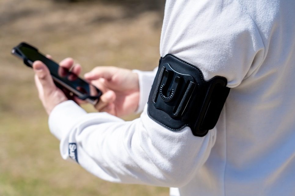 A person in a white sweatshirt holds a phone in both hands while a black band with an empty mount stays on the upper arm.