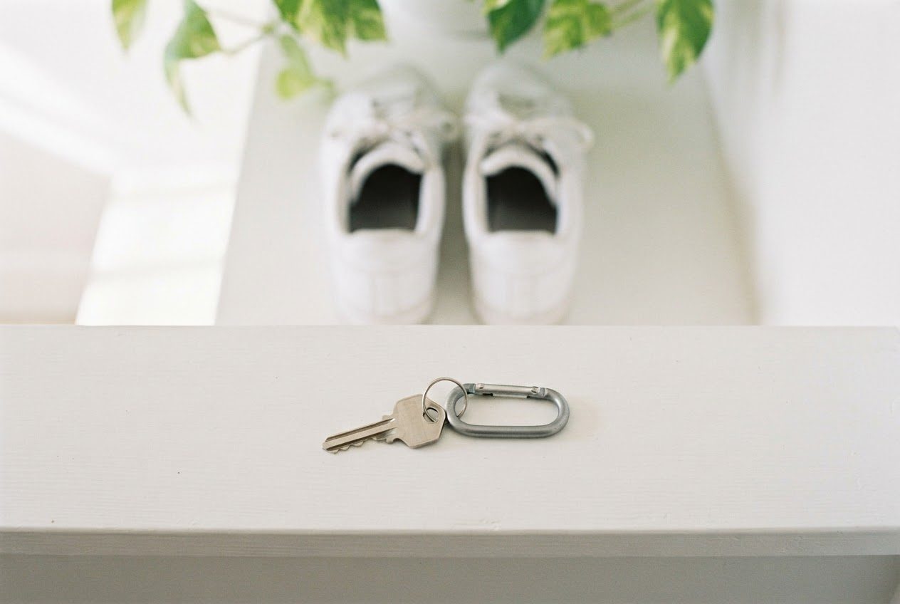 Close view of a single house key clipped to a small carabiner resting on a hallway shelf, morning light, running shoes blurred behind