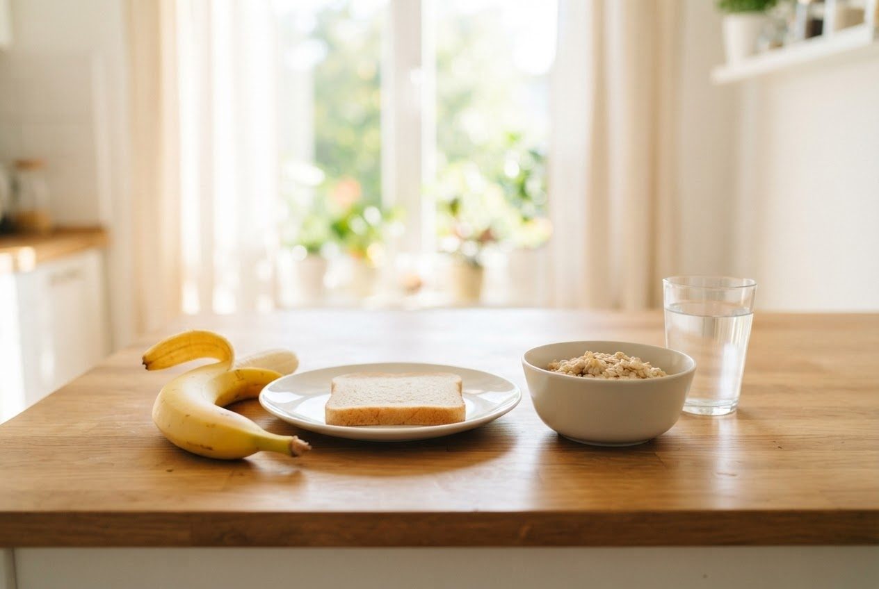 A small pre-run snack on a bright sunlit kitchen table: one banana, a single slice of plain toast, a small bowl of oatmeal and a glass of water — a light snack portion, clearly not